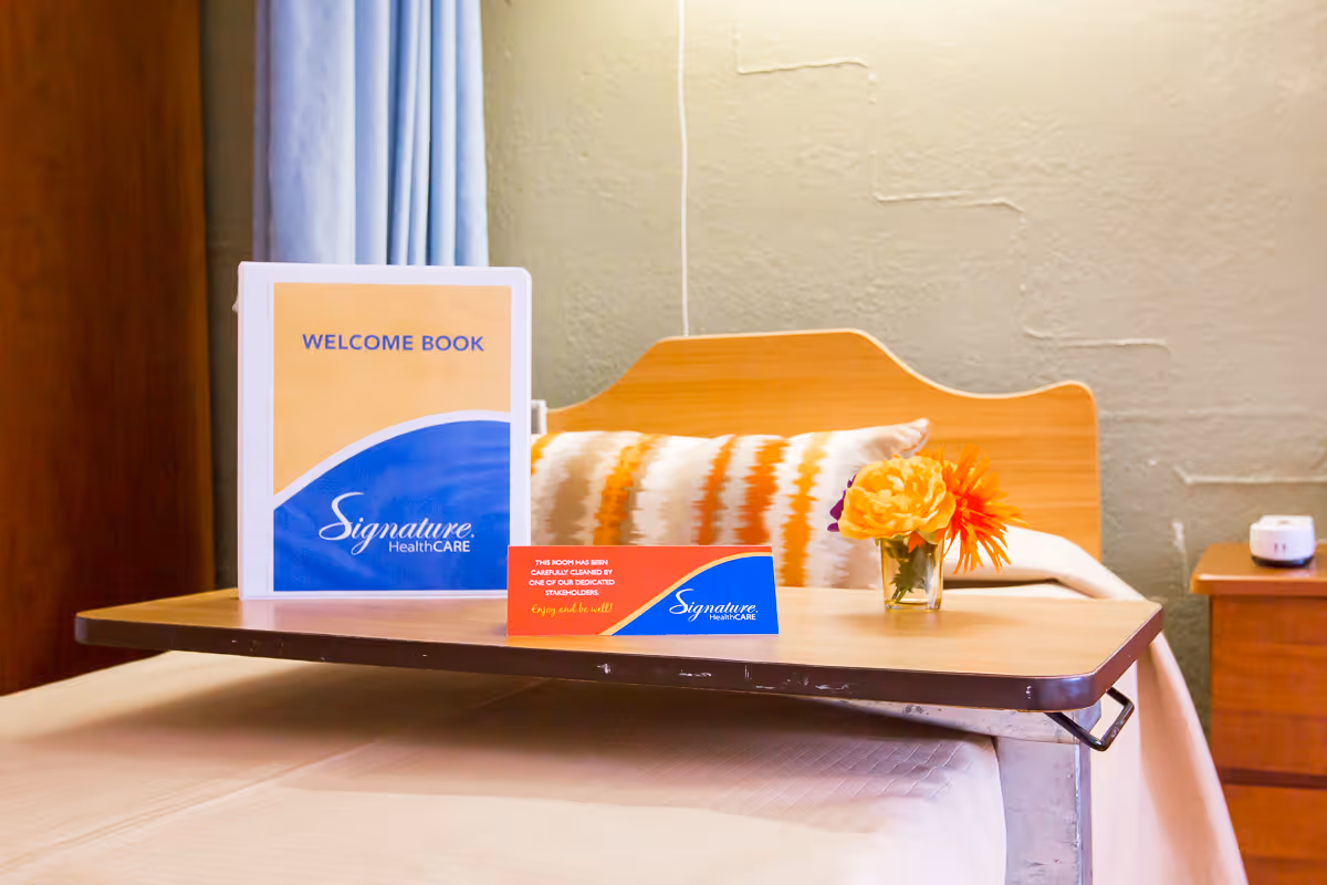 A hospital-style bed with a wooden headboard and a patterned pillow. On the bed is a wooden overbed table holding a welcome book labeled Signature HealthCARE, a small card indicating the room has been cleaned, and a small vase with orange and yellow flowers. A wooden nightstand with a small white device is visible in the background.