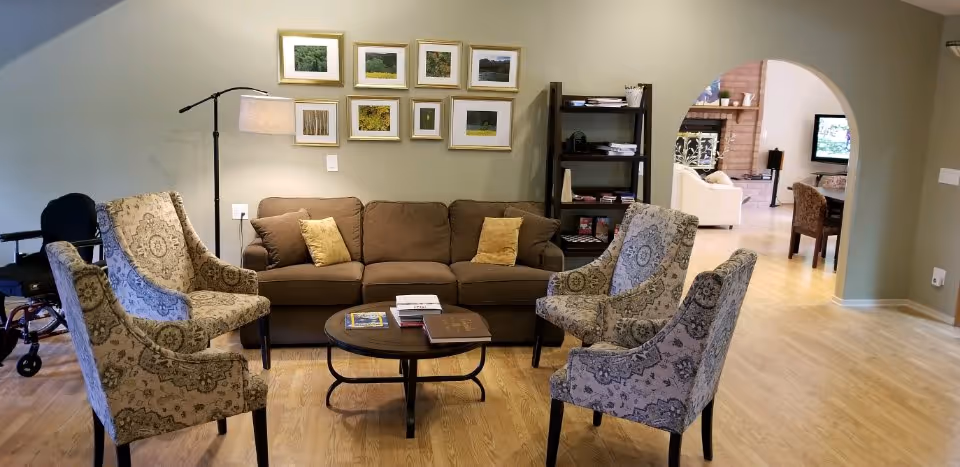 A cozy living room area with a brown sofa, four patterned armchairs arranged around a round wooden coffee table with books on it. A floor lamp stands beside the sofa, and a set of framed pictures hangs on the wall above it. In the background, an open archway leads to another room with a fireplace, TV, and additional seating. A wheelchair is partially visible on the left side of the image.