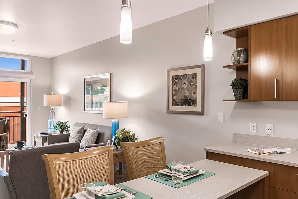 Interior view of a senior living facility showing a dining area with a table set for two, adjacent to a living room with a gray sofa, side tables with lamps, and framed artwork on the walls. The kitchen area with wooden cabinets and a countertop is partially visible.