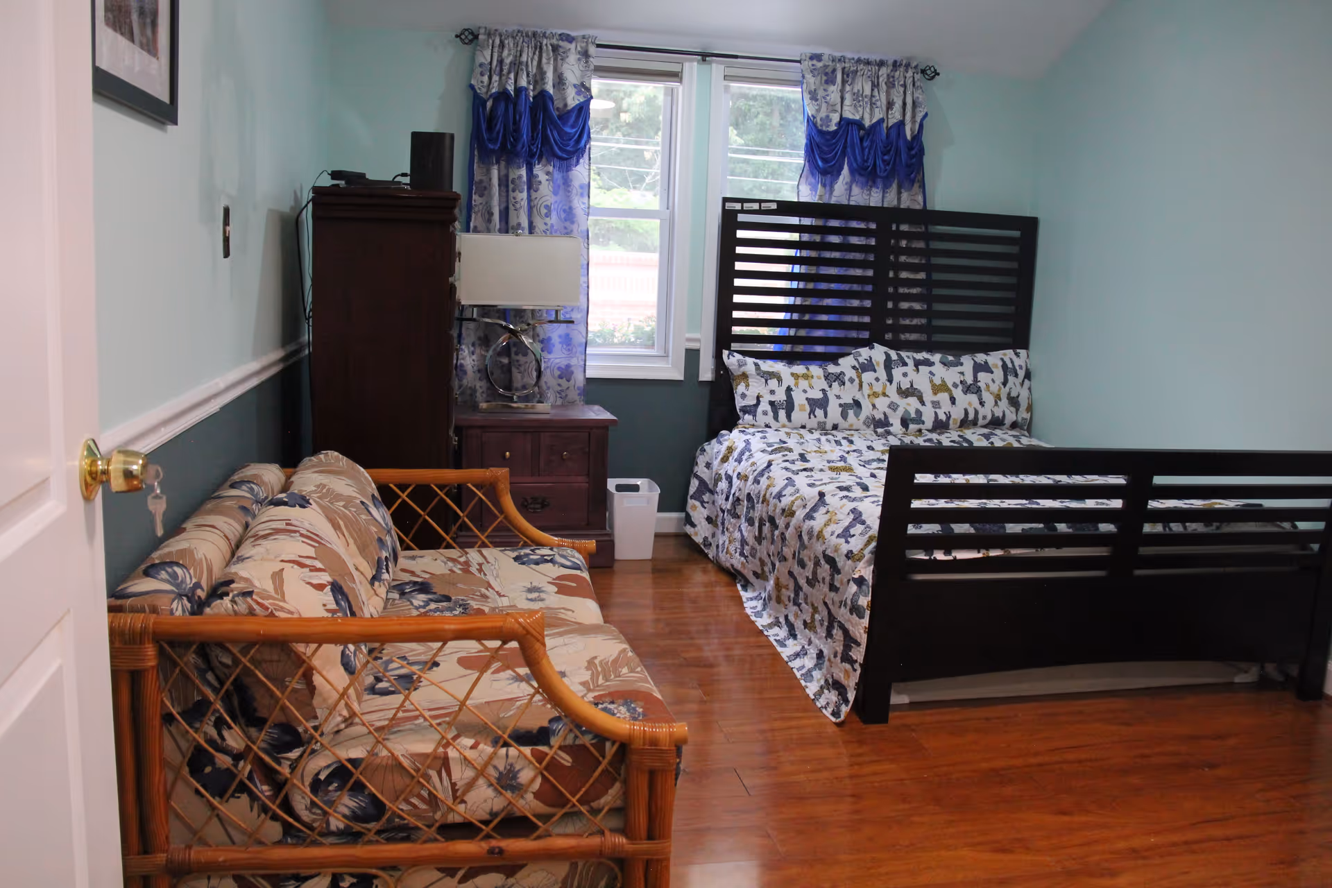 A bedroom with light blue walls and wooden floor. The room features a dark wooden bed with a colorful animal-patterned bedspread and matching pillows. Next to the bed is a wooden nightstand with a modern lamp and a tall dresser. A floral-patterned sofa with a wooden frame is positioned along the left wall. Two windows with blue and white curtains allow natural light into the room.
