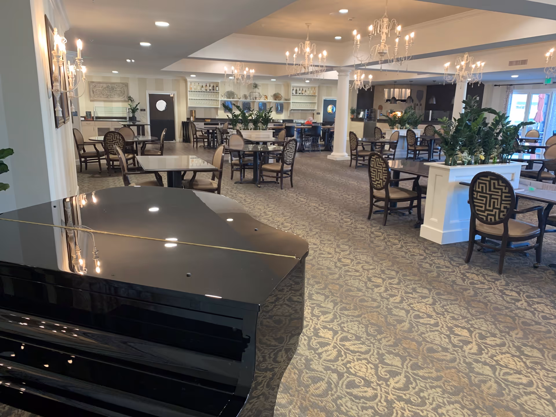 Spacious dining area with multiple tables and chairs arranged neatly. A black grand piano is visible in the foreground on the left. The room features chandeliers hanging from the ceiling, patterned carpet flooring, and plants placed on white dividers. Large windows and glass doors allow natural light to enter the room.