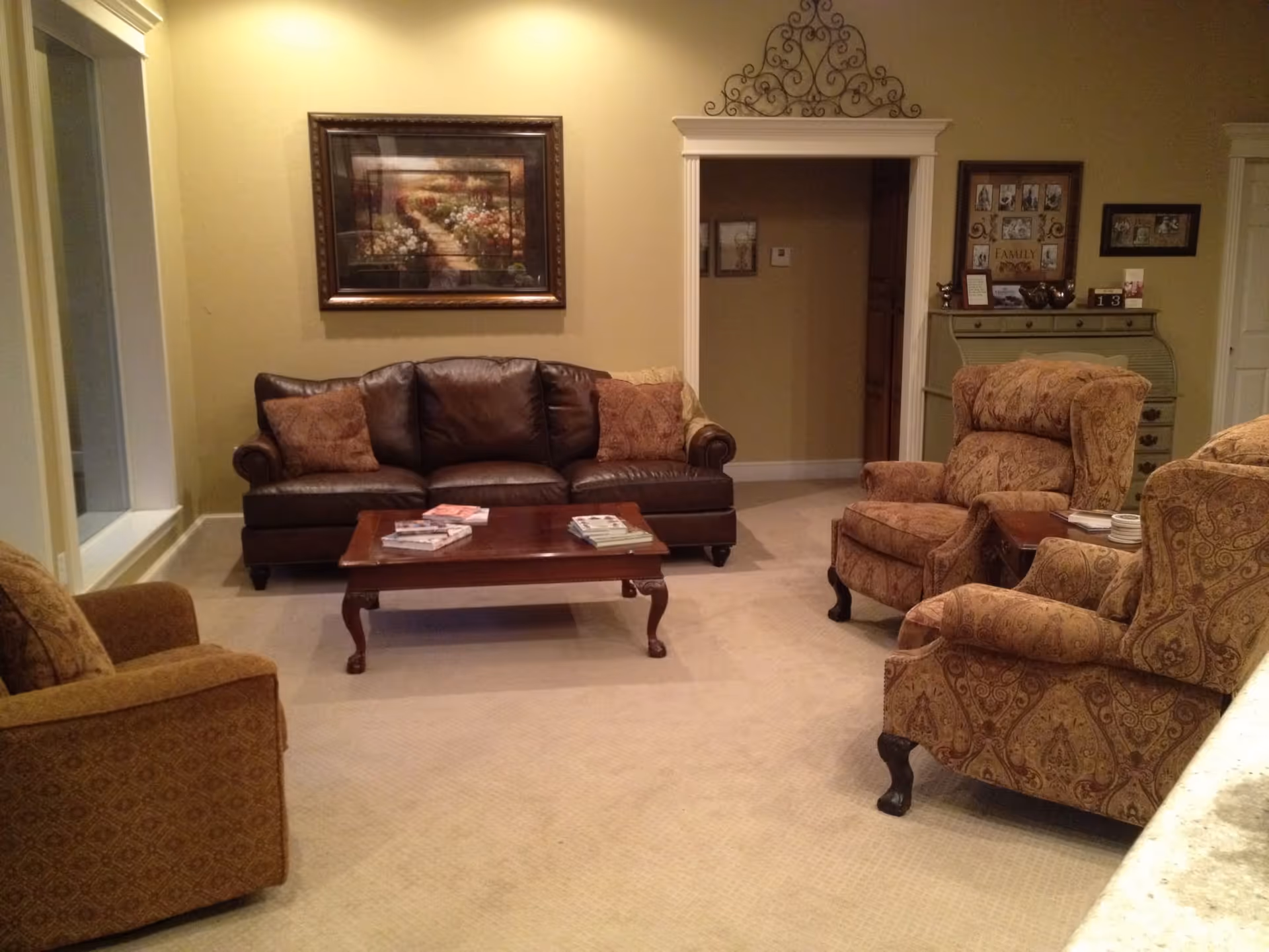 A cozy living room with a brown leather sofa, three patterned armchairs, a wooden coffee table with books on it, beige carpet, and a framed painting of a garden pathway on the wall. There is a decorative metal wall piece above a doorway and a greenish chest with family photos and decor on top.