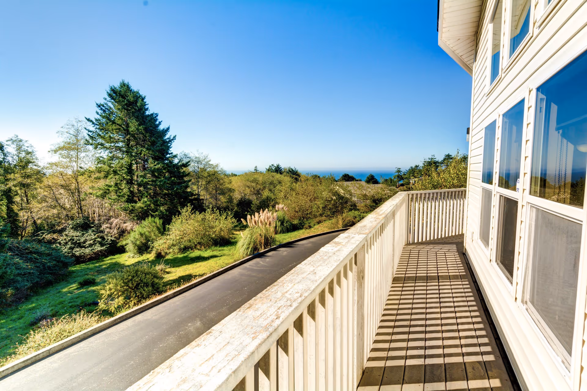 View from a wooden balcony with a white railing and windows, overlooking trees and the distant ocean under a clear blue sky.