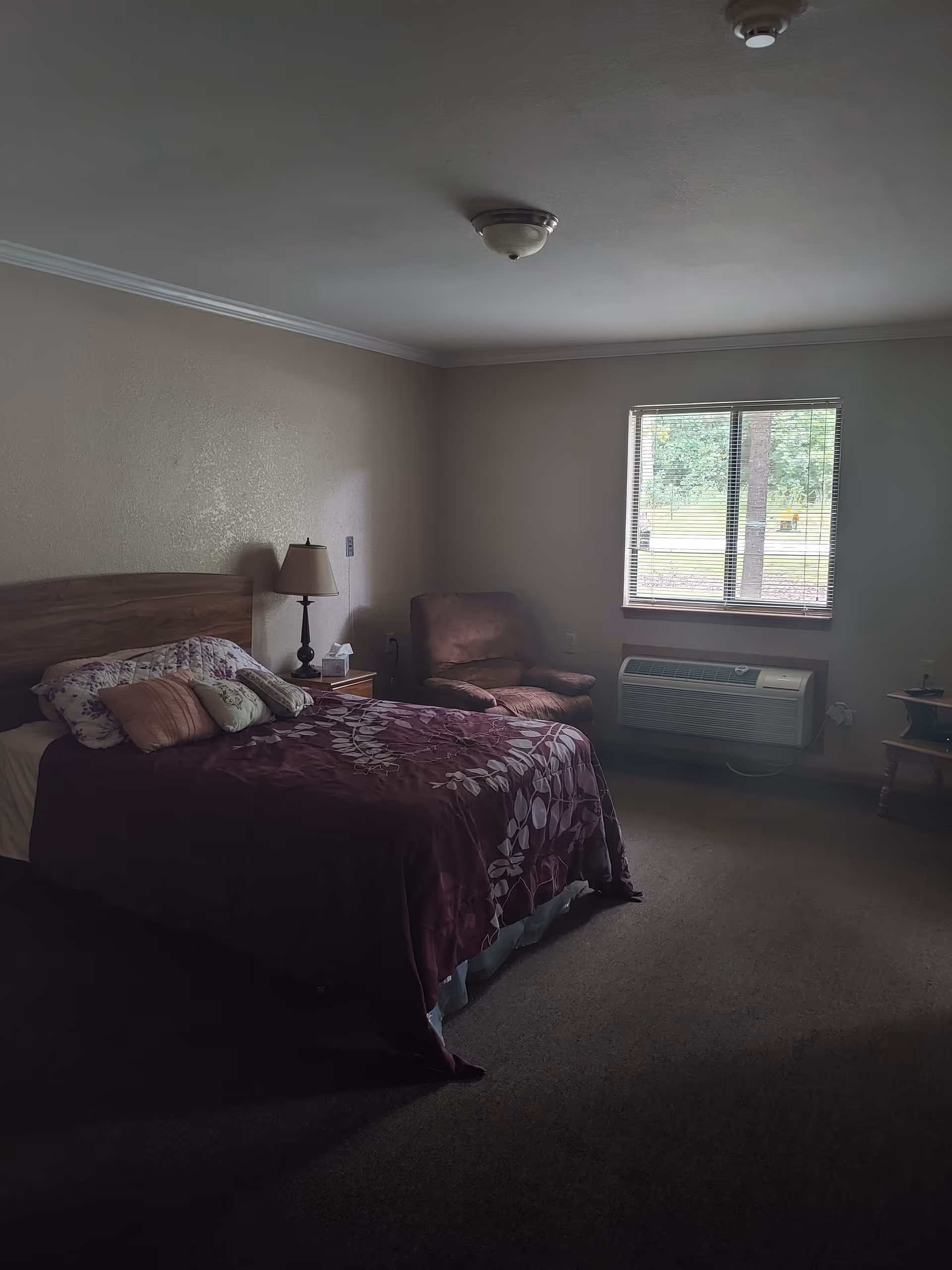 A bedroom with a bed covered in a purple floral bedspread and several pillows. Next to the bed is a wooden nightstand with a lamp and a box of tissues. There is a cushioned armchair near a window with blinds, and below the window is a wall-mounted air conditioning unit. The room has beige walls and carpeted floor.