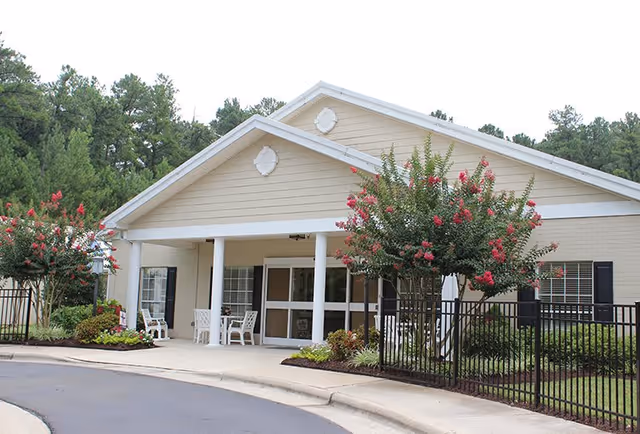 Exterior view of a single-story beige building with white trim and columns at the entrance. There are flowering trees with pink blossoms and a black metal fence surrounding the landscaped area. White chairs and a small table are placed near the entrance under the covered porch.