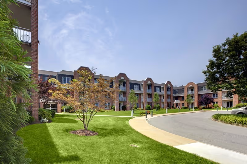 Exterior view of a multi-story senior living facility building with brick facade, balconies, and well-maintained green lawn and trees under a clear blue sky.