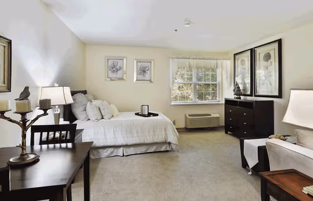 A bright and neatly arranged senior living bedroom with a bed covered in white linens, a dark wooden dresser with decorative items, framed artwork on the walls, a window with sheer curtains, a small table with a lamp and candles, and a comfortable chair.