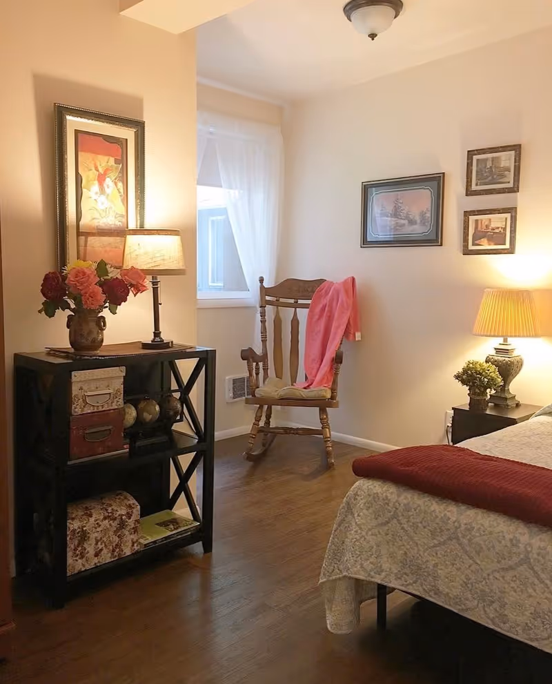 A cozy bedroom corner with a wooden rocking chair draped with a pink blanket, a small black shelf holding decorative boxes and a vase with flowers, a table lamp, framed artwork on the walls, and part of a bed with a patterned bedspread and a red throw blanket.
