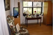 A cozy room with a floral upholstered armchair, a wooden cabinet with a flat-screen TV on top, and a wooden desk by a large window with plants and decorative items. The room has light-colored wooden flooring and neutral walls.