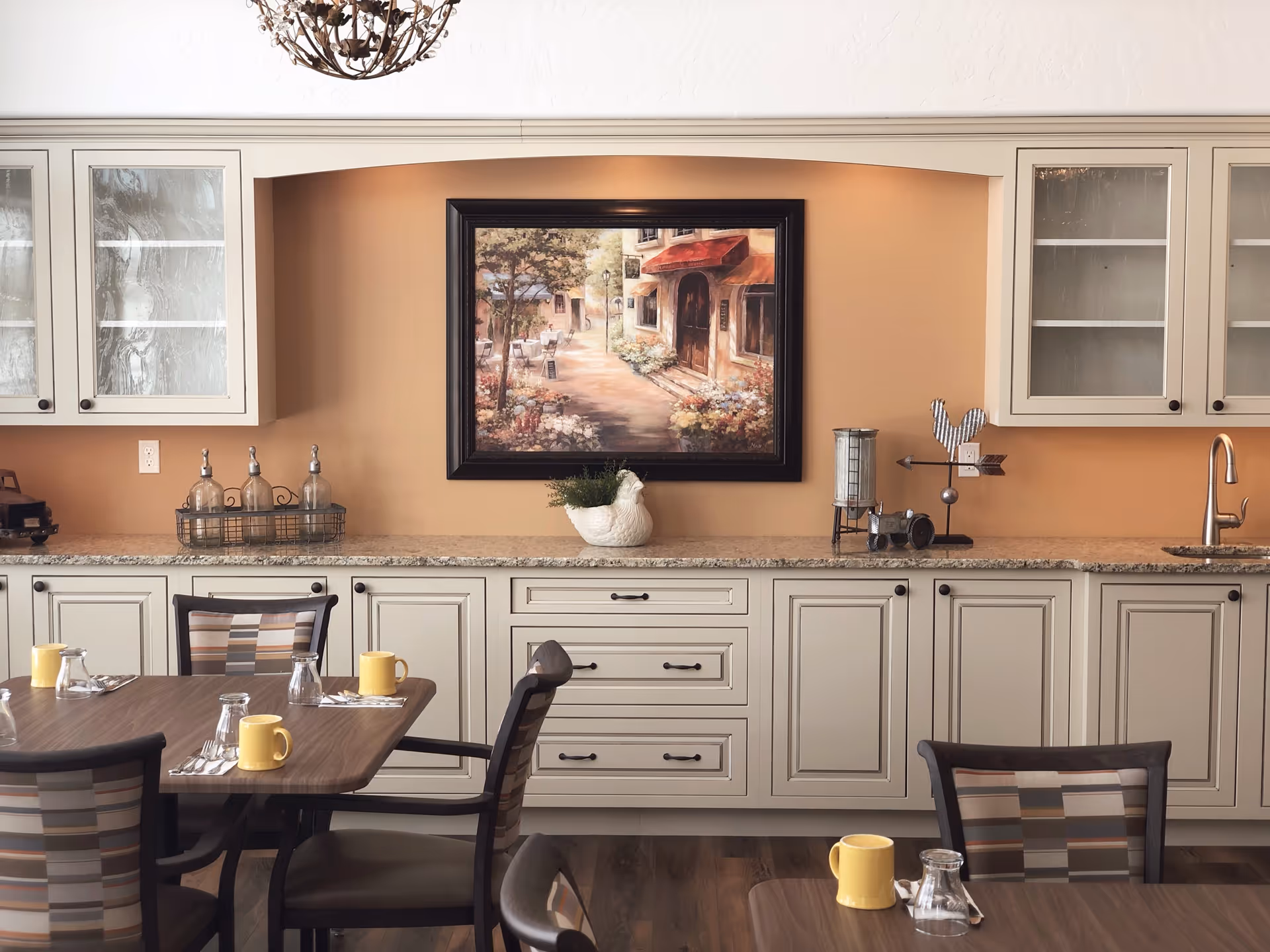 A dining area with a wooden table set with yellow mugs, glasses, and silverware. The background features beige cabinets with glass doors, a granite countertop, a small sink, and decorative items including a framed painting of a street scene, a plant in a white pot, and a metal rooster figurine.