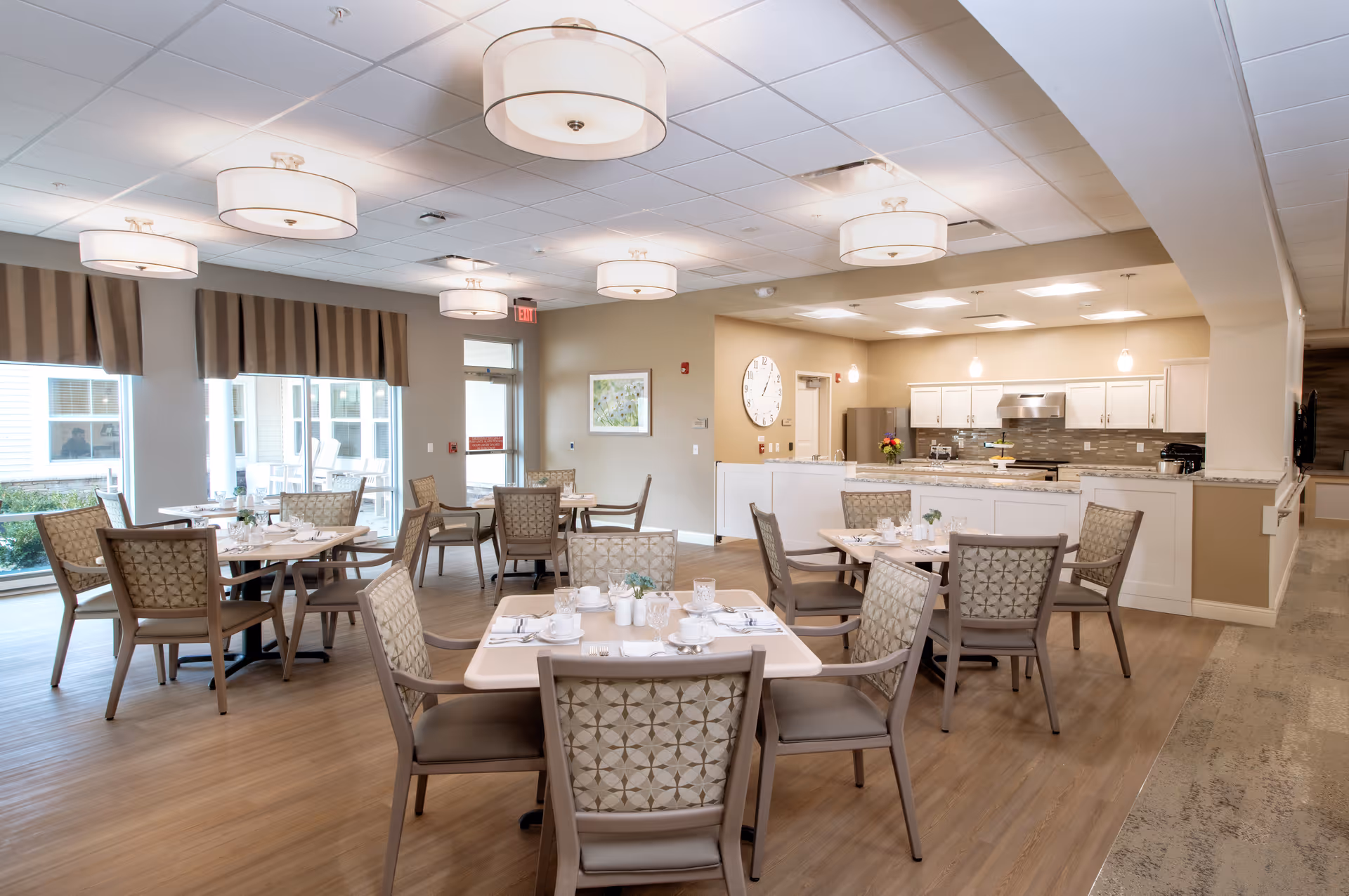 A bright and spacious dining room with several tables and chairs arranged neatly. Each table is set with white dishes, cups, and silverware. The room features large windows with striped valances allowing natural light to fill the space. In the background, there is a modern kitchen area with white cabinets, a stainless steel refrigerator, and a large clock on the wall. The ceiling has multiple round light fixtures.