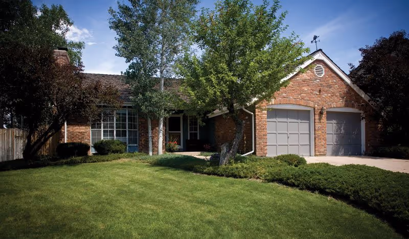 Front exterior view of a single-story brick house with a well-maintained lawn, several trees, and a two-car garage with closed gray doors under a clear blue sky.