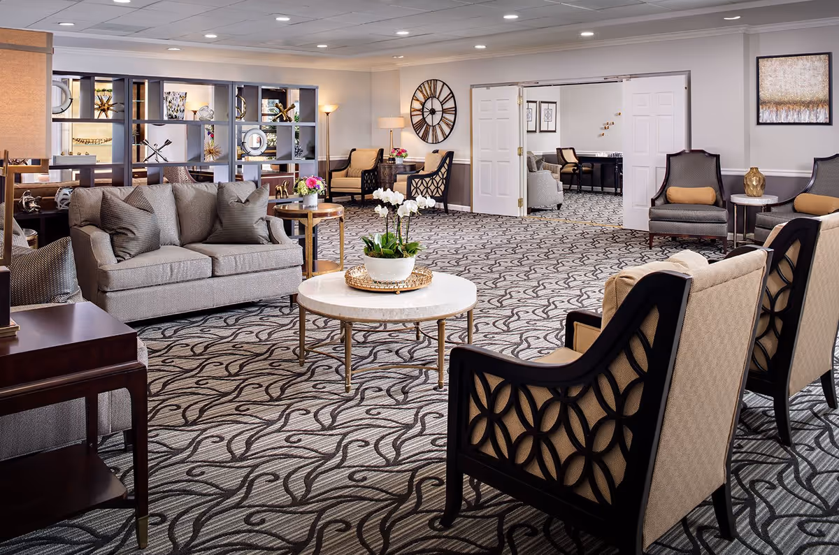 A spacious and elegantly furnished senior living facility common area with patterned carpet, beige sofas, armchairs, a round marble coffee table with a white orchid centerpiece, decorative shelving, and wall art. The room is well-lit with ceiling lights and lamps, and there is an adjoining room visible through open double doors.