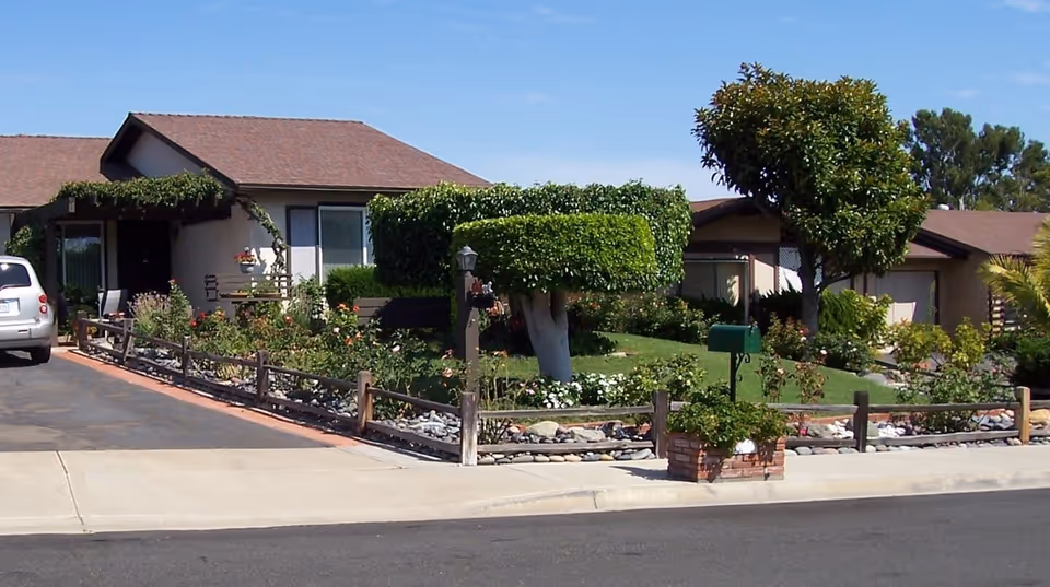 Exterior view of a single-story residential building with a brown roof and beige walls. The front yard features neatly trimmed bushes, a small tree, a green mailbox, and a garden with various plants and flowers. A white vehicle is parked on the driveway to the left.