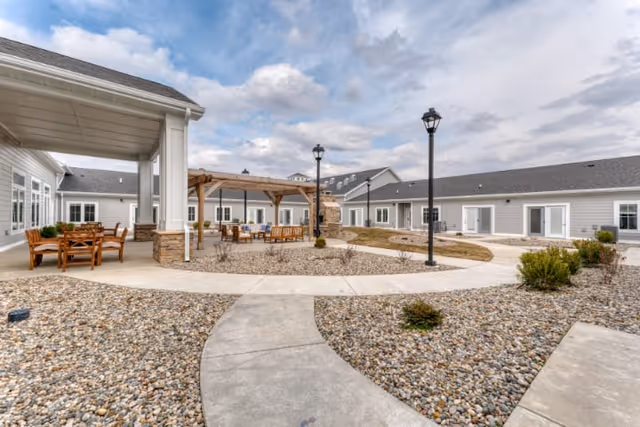 Sunny courtyard with paved walkways, a wooden pergola and outdoor seating surrounded by single-story senior living buildings.