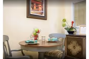 A small round wooden dining table set for two with teal napkins and a centerpiece of green apples in a glass bowl. Two chairs with gray backs and striped cushions are positioned around the table. A framed painting hangs on the beige wall above the table, and a decorative cabinet with a plant and framed certificate is visible to the right.