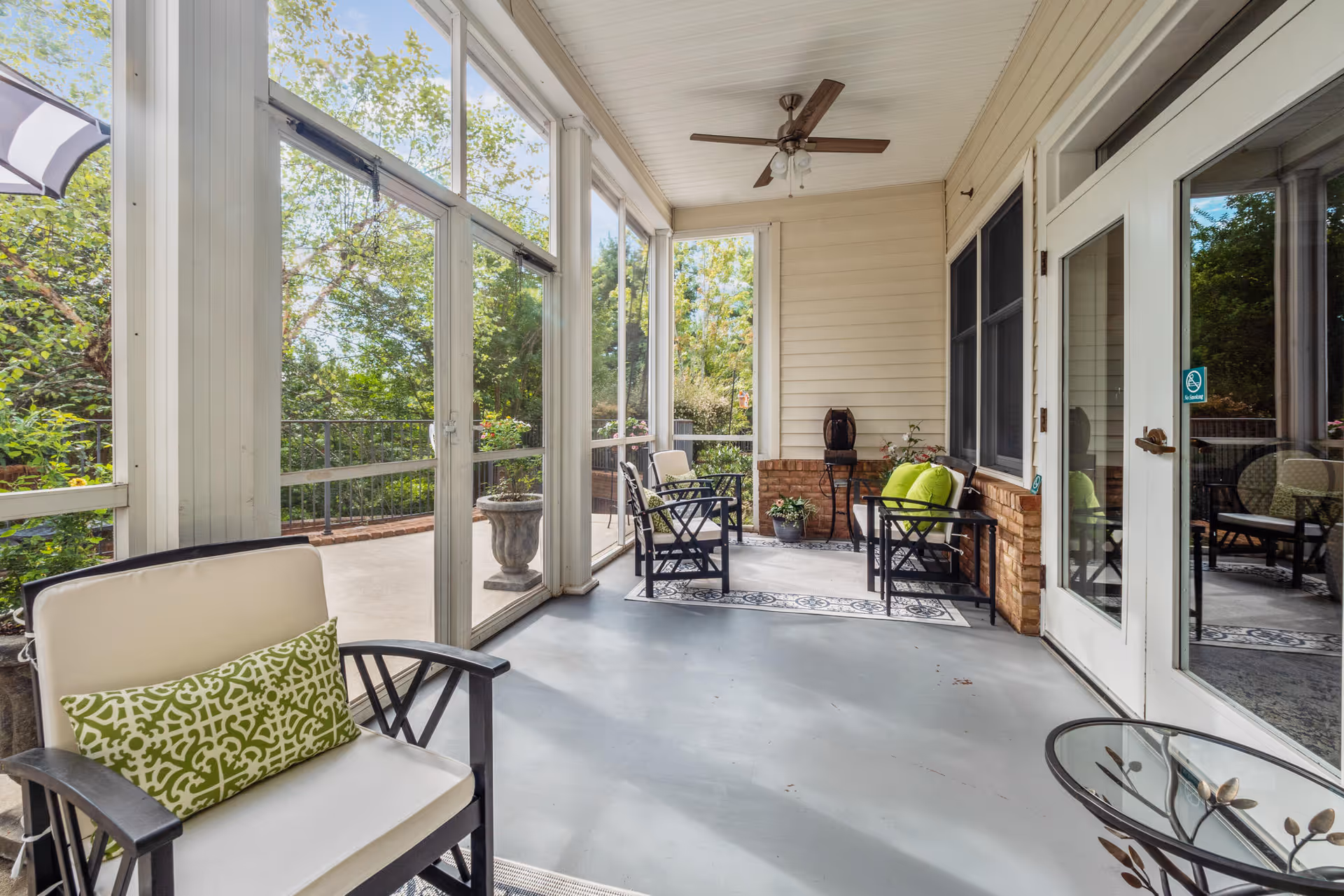 A bright screened-in porch area with several cushioned chairs, green decorative pillows, a ceiling fan, potted plants, and large windows providing a view of trees and greenery outside.