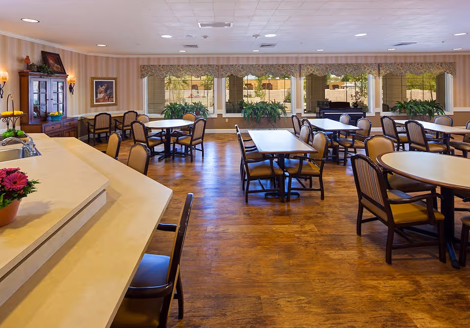 A spacious dining room with multiple tables and chairs arranged neatly. The room features large windows with floral valances, allowing natural light to brighten the space. There are several potted plants on the windowsills and a wooden cabinet with decorative items on the left side. The floor is wooden, and the ceiling has recessed lighting.