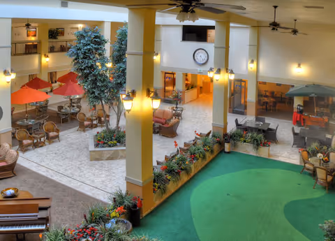Indoor atrium area of a senior living facility with seating arrangements including wicker chairs and tables with red and green umbrellas. There are large potted plants, ceiling fans, and a clock mounted on the wall above a reception or common area. The floor is tiled with a section of green carpet resembling a putting green, and the space is well-lit with wall-mounted lamps.