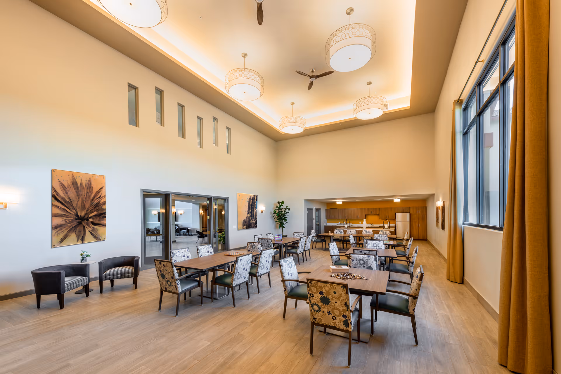 A spacious dining room with multiple tables and chairs arranged neatly. The room features high ceilings with modern light fixtures and ceiling fans. Large windows with mustard-colored curtains allow natural light to fill the space. The back of the room has a kitchen area with wooden cabinets and appliances. Two armchairs and wall art decorate one side of the room.
