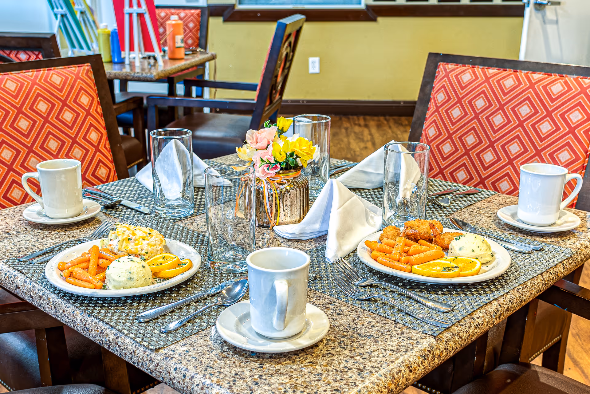 Dining table set for four with plates of food, coffee cups, glasses, napkins and a small flower centerpiece in a dining room.