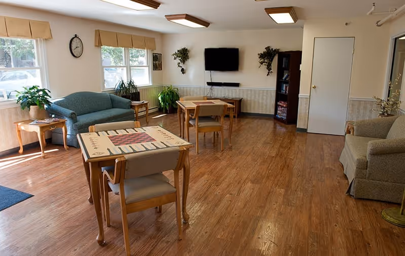 A bright communal lounge with wood floors, game tables and chairs, sofas, plants, and a wall-mounted TV.