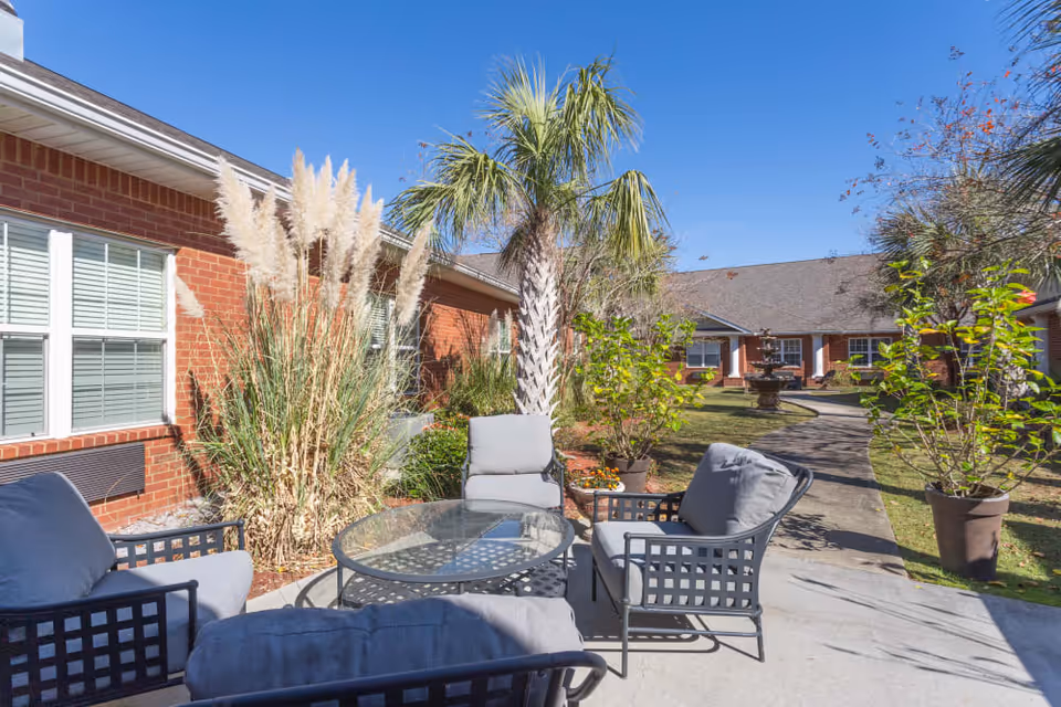 Sunny courtyard patio with cushioned outdoor seating and a glass-top table in front of a brick senior living building.
