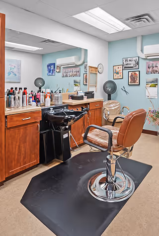 Interior view of a hair salon area with a brown salon chair on a black mat in front of a wooden counter with various hair care products. There is a black hair washing sink attached to the counter. The walls are light blue and decorated with framed pictures and a calendar. A hair dryer hood is mounted on the wall.