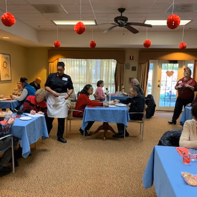 A dining room in a senior living facility with several elderly residents seated at tables covered with blue tablecloths. A staff member in a black shirt and white apron is standing and interacting with the residents. Red hanging decorations are suspended from the ceiling, and large windows with curtains allow natural light into the room.