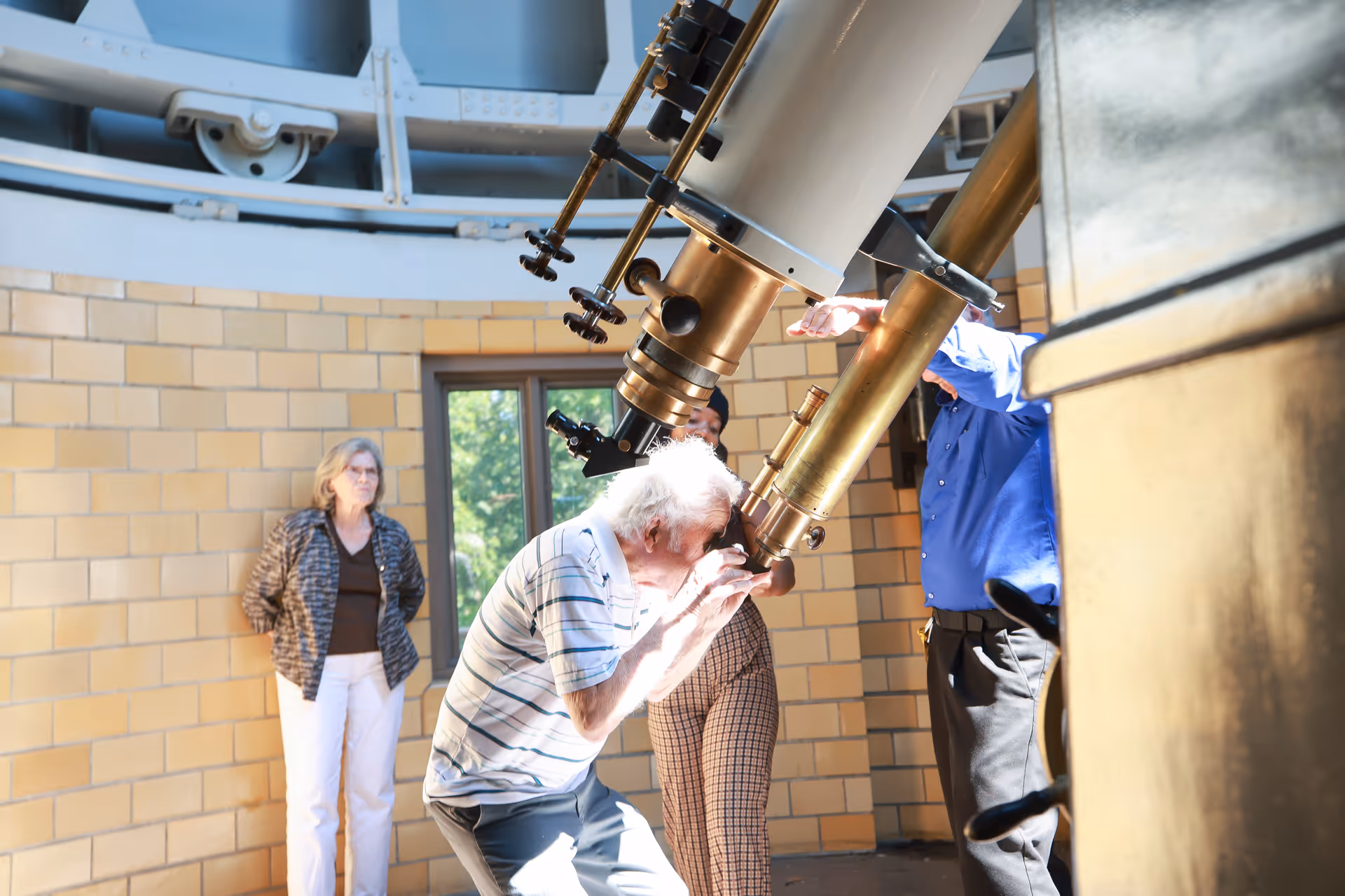 An elderly man looking through a large telescope inside an observatory dome, accompanied by two other adults watching nearby. The room has beige brick walls and a window showing greenery outside.