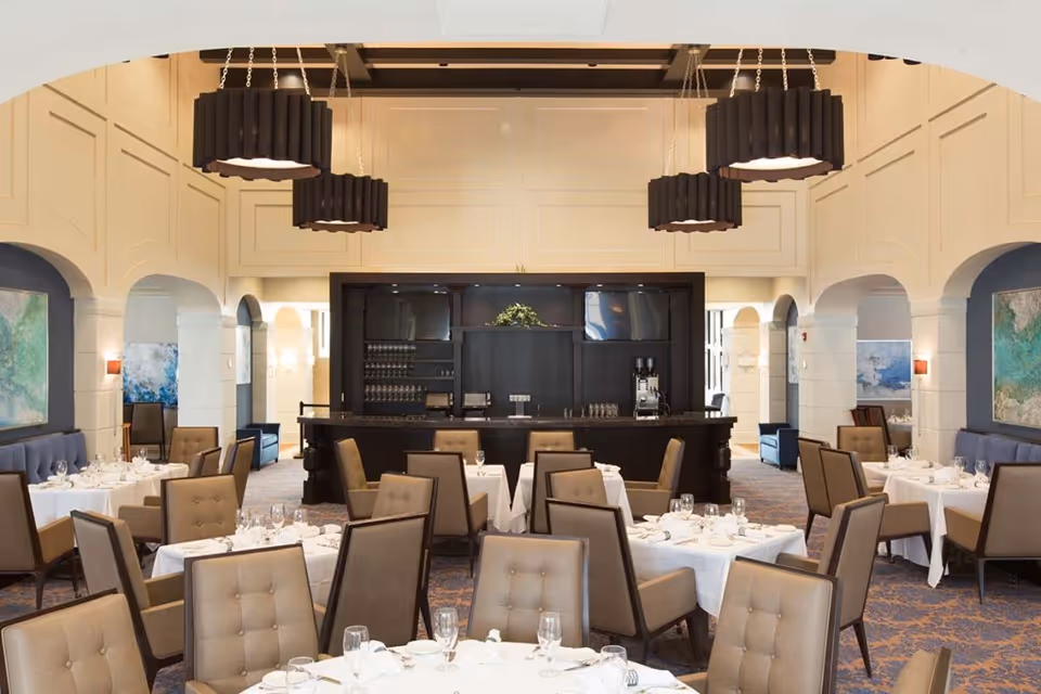 Elegant dining room with multiple tables covered in white tablecloths, set with glassware and napkins. The room features beige upholstered chairs, a dark wooden bar in the background, large modern pendant lights hanging from the ceiling, and decorative artwork on the walls.