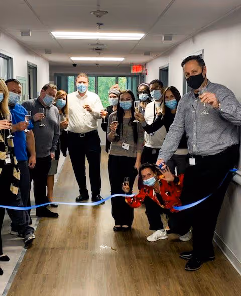 A group of people standing in a hallway, wearing face masks and holding glasses, gathered around a blue ribbon for a ribbon-cutting ceremony at Majestic Oaks Rehabilitation and Nursing Center.
