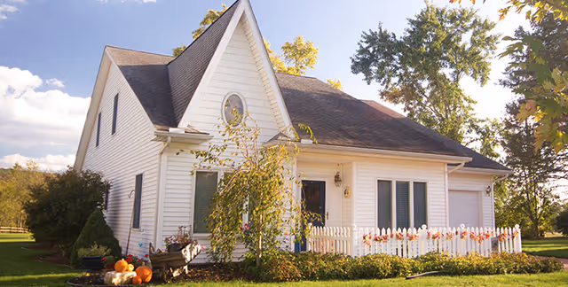A white house with a steep roof and an oval window near the peak, surrounded by green grass and trees under a partly cloudy sky. The house has a small white picket fence decorated with autumn-themed ornaments and pumpkins placed near the front yard.