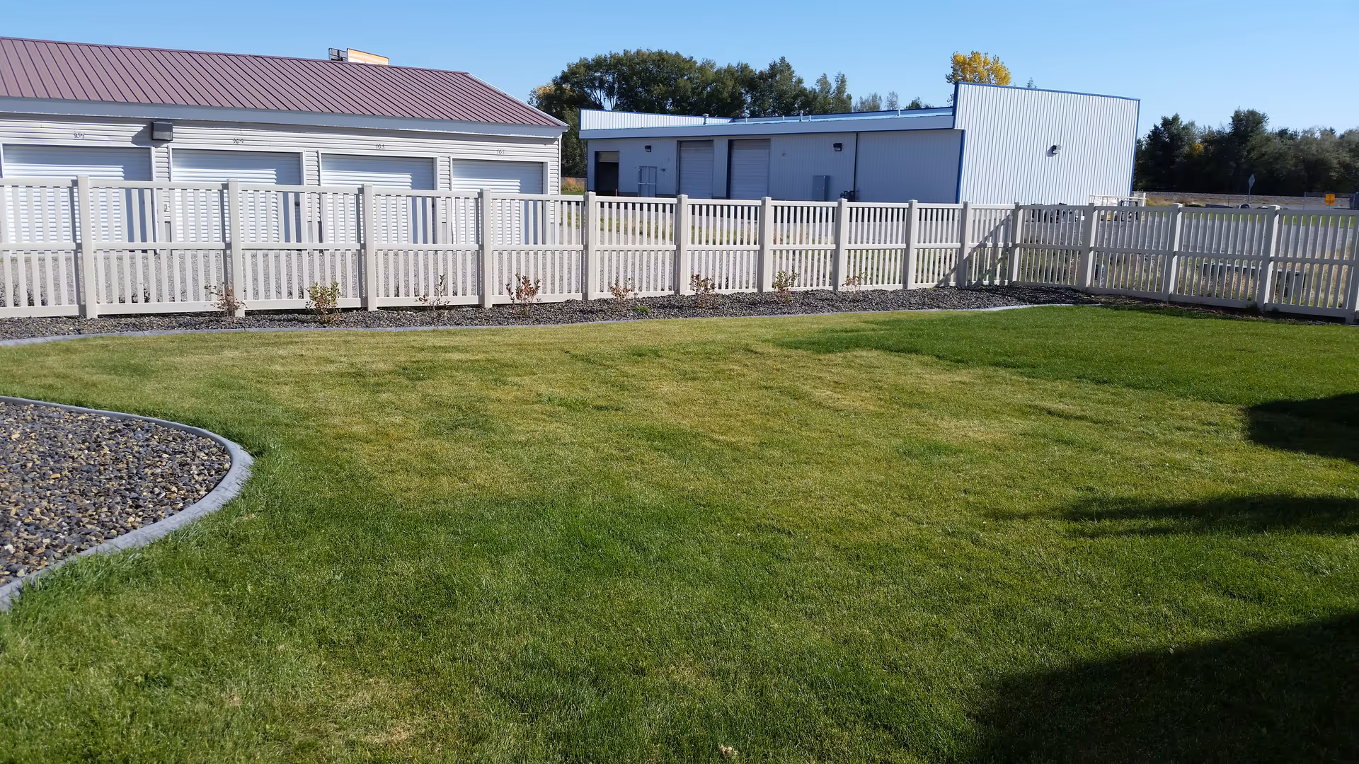 A fenced outdoor grassy area with a white picket fence surrounding it. There are two buildings in the background, one with a red roof and the other with a white exterior. The sky is clear and blue with some trees visible behind the buildings.