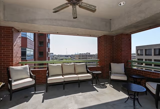 Covered outdoor patio area with cushioned seating including a three-seat sofa and three armchairs, small round tables, brick pillars, and a ceiling fan. The patio overlooks nearby buildings and a distant cityscape under a clear sky.