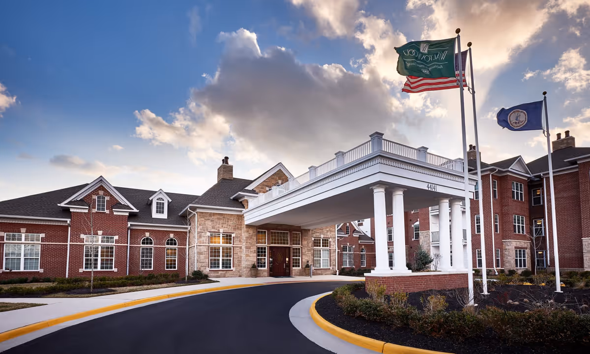 Entrance of a brick senior living facility with a covered porte-cochere, three flagpoles, and a landscaped circular driveway under a cloudy sky.