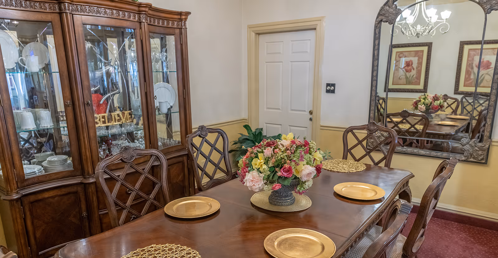 Formal dining room with a wooden table set with gold chargers and a floral centerpiece, a china cabinet and large mirror on the wall.