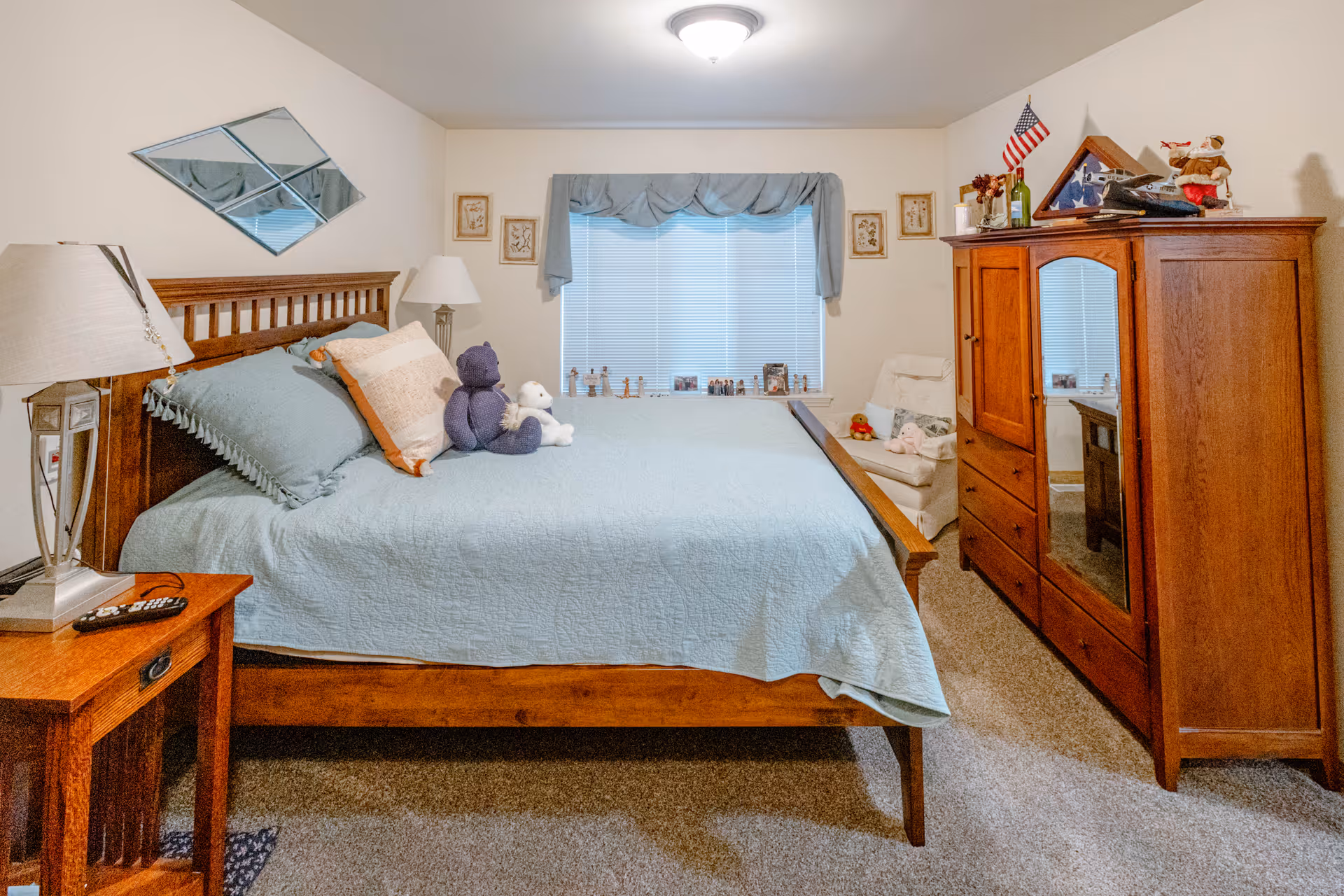 A cozy bedroom with a wooden bed frame and a light blue quilt. Two stuffed animals, a purple bear and a white bear, sit on the bed. There is a wooden nightstand with a lamp and a remote control on it. A wooden wardrobe with a mirror on one door stands to the right, topped with decorative items including a small American flag. A window with blue curtains is centered on the back wall, flanked by framed pictures. A white armchair with a pillow and stuffed toys is positioned near the wardrobe. The room has beige walls and carpeted floor.
