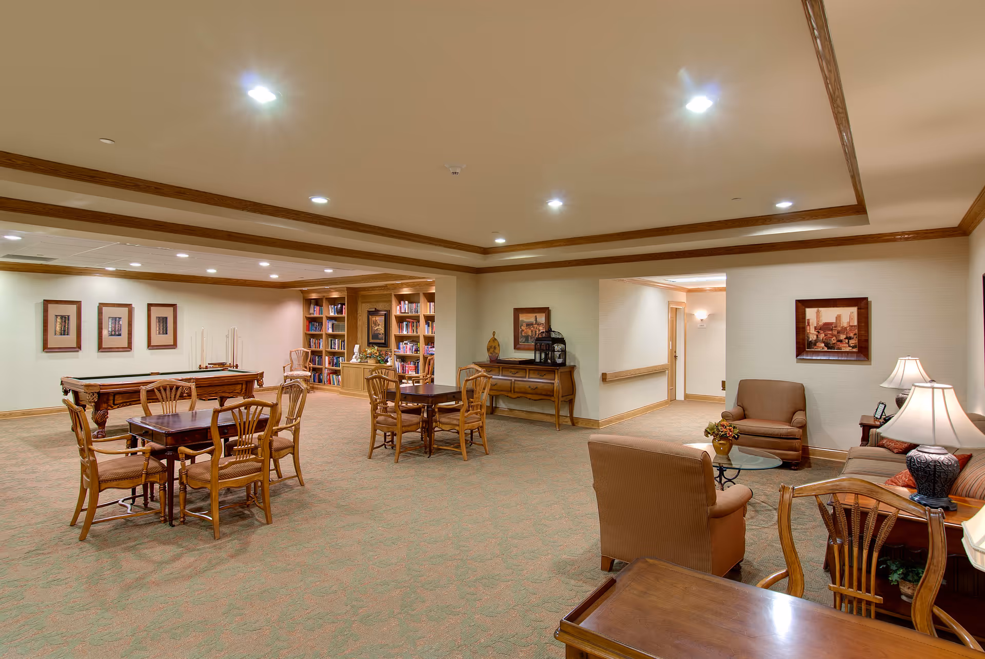 A spacious, well-lit common area with multiple wooden tables and chairs, a pool table, bookshelves filled with books, and comfortable seating including armchairs and a sofa. The room has beige walls, carpeted floors, framed artwork on the walls, and decorative lamps providing warm lighting.