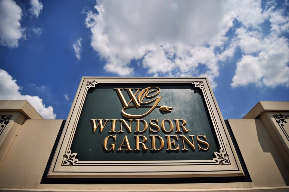 Ornate green and gold 'Windsor Gardens' entrance sign set against a blue sky with clouds.