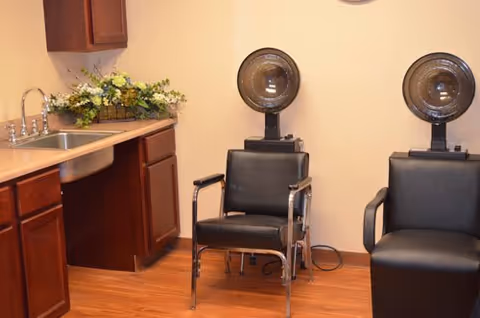 Two black salon chairs with hooded hair dryers next to a sink, wooden cabinets, and a floral arrangement.