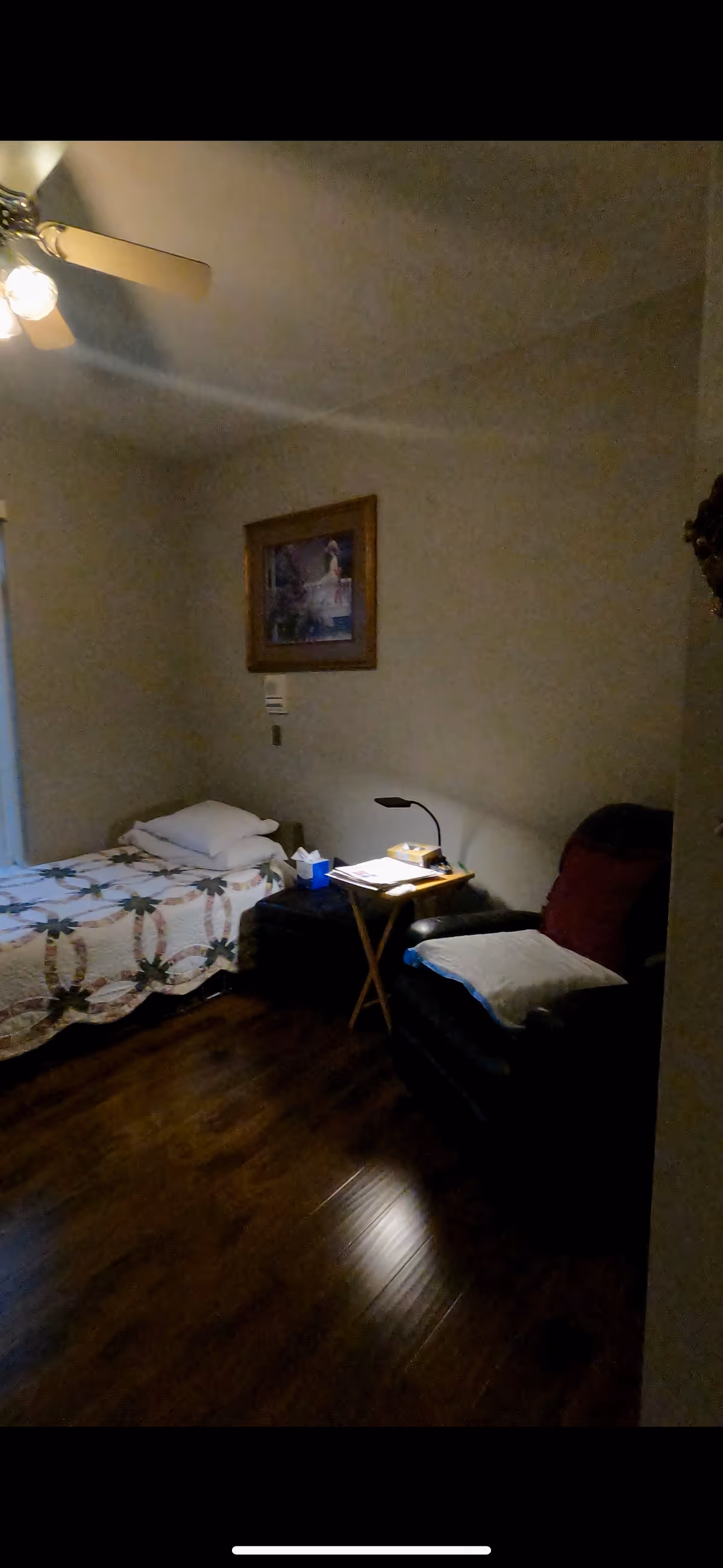 A small bedroom with a single bed covered with a patterned quilt, a pillow, and folded linens. Next to the bed is a small wooden side table with a lamp, a box of tissues, and some papers. There is a dark armchair with a cushion and a red throw pillow. A framed picture hangs on the wall above the bed. The room has wooden flooring and a ceiling fan with lights.