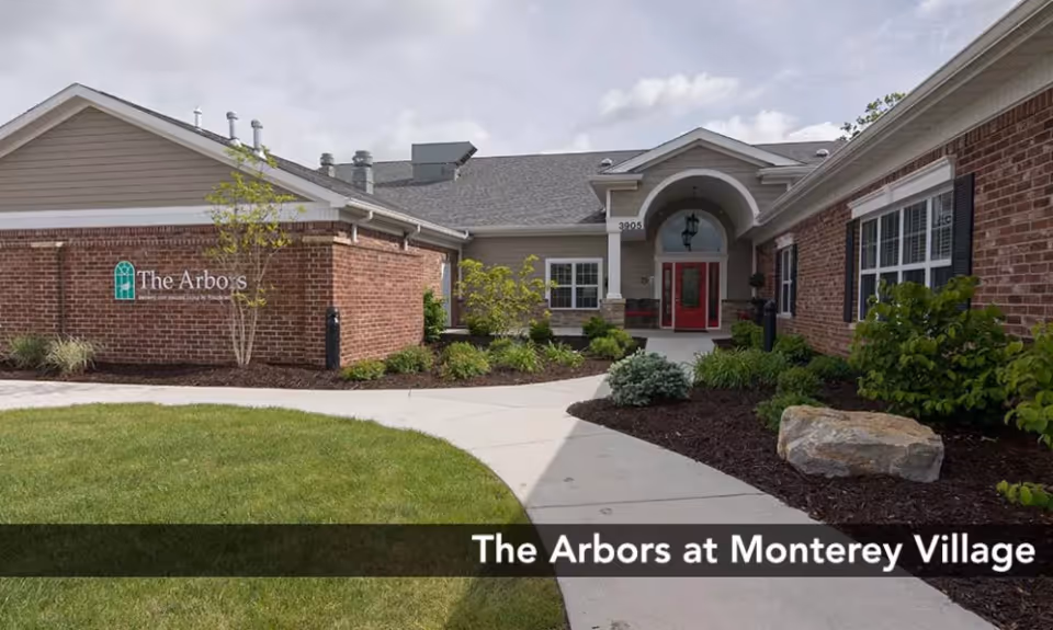Front entrance of a single-story brick senior living building with a paved walkway, landscaping, and a sign for The Arbors.