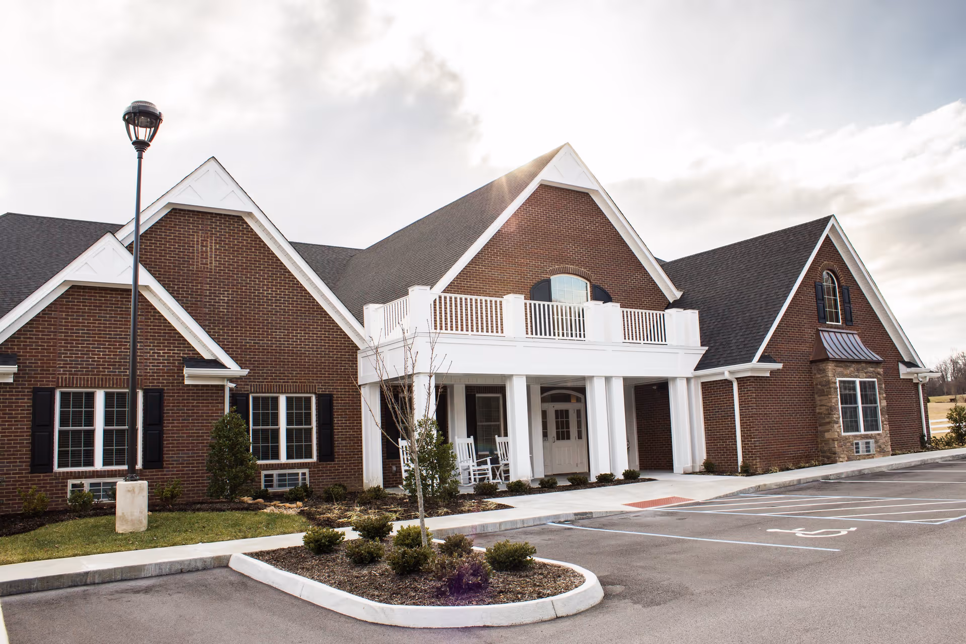 Exterior view of a senior living facility building with brick walls, white trim, and a covered entrance with white columns. There are rocking chairs on the porch, a lamp post, landscaped bushes, and a parking lot with marked spaces including a handicapped spot.
