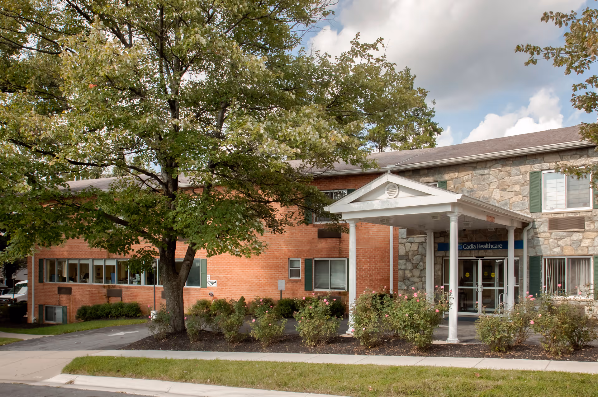 Exterior view of a two-story senior living facility with a brick and stone facade, a covered entrance supported by white columns, surrounded by greenery and a large tree in front.