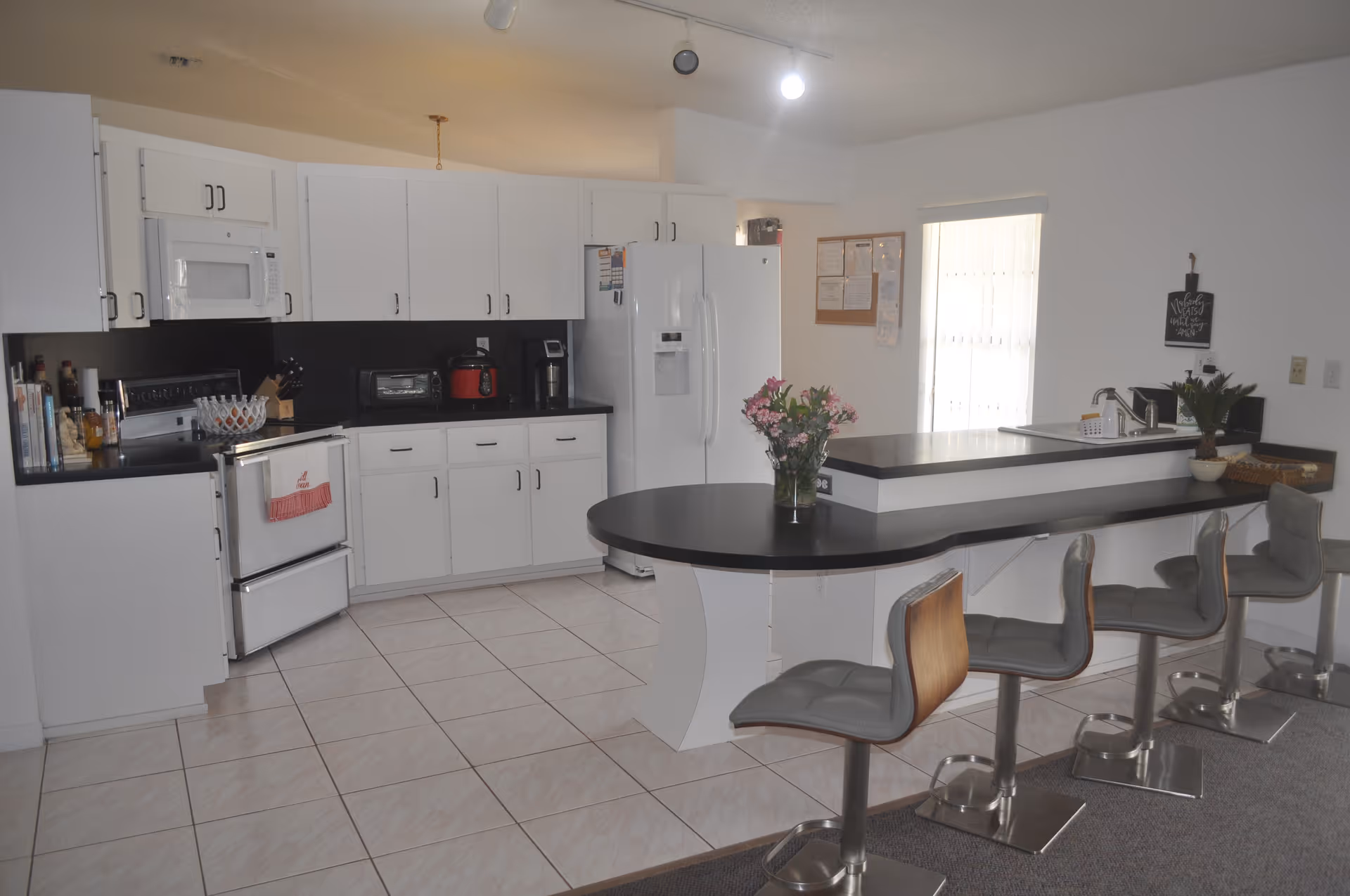 A modern kitchen with white cabinets, a white refrigerator, a stove, microwave, and various kitchen appliances on the black countertops. There is a curved black countertop island with four gray bar stools, a vase with flowers on the island, and a window with blinds letting in natural light.