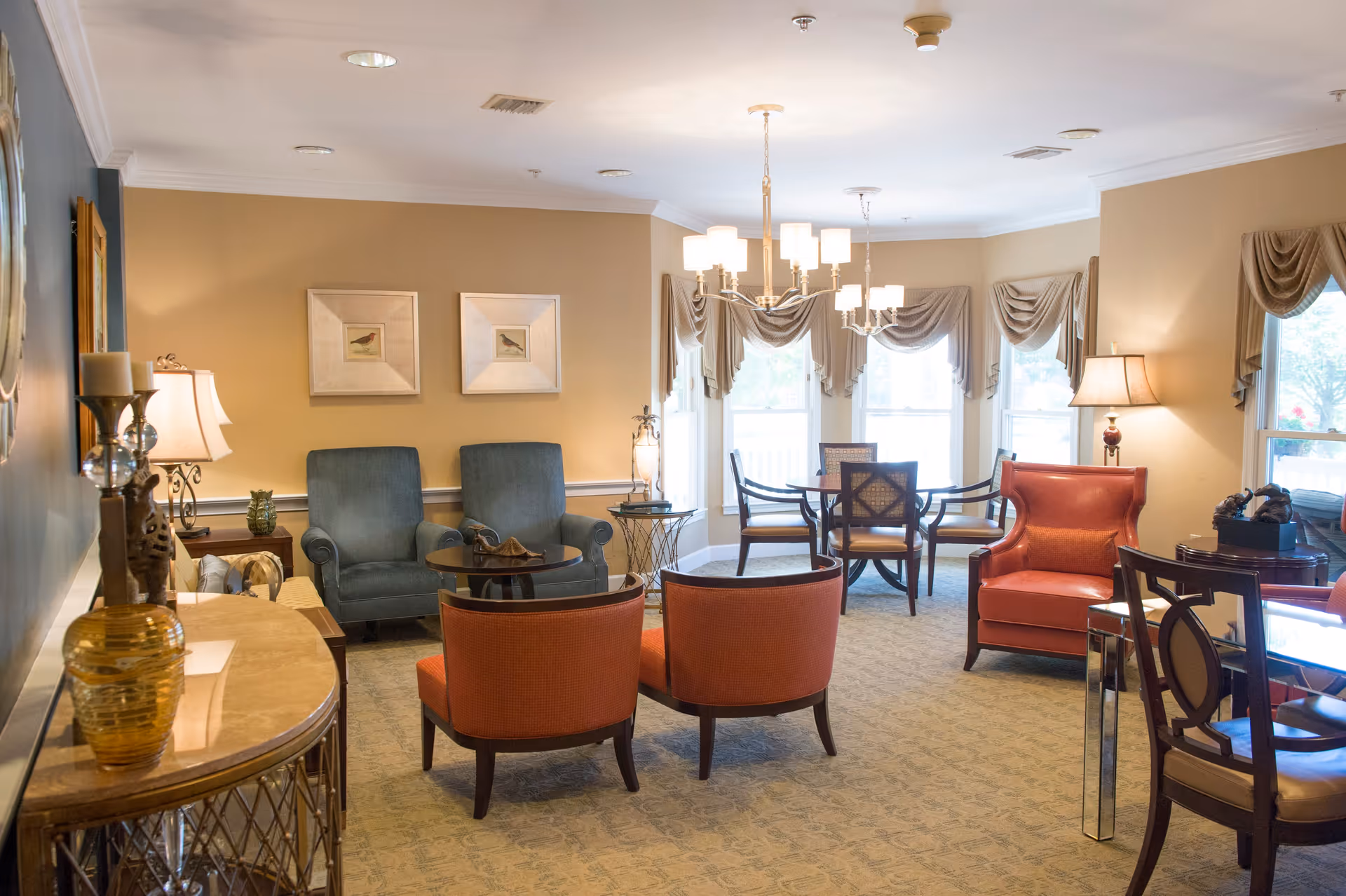 A well-lit living room area with beige walls and carpeted floor. The room features multiple seating options including two blue armchairs, two orange armchairs, a red armchair, and several wooden chairs around a glass table and a round dining table near windows with draped curtains. There are two chandeliers hanging from the ceiling and several lamps providing additional lighting. Decorative items and framed pictures adorn the walls and furniture.