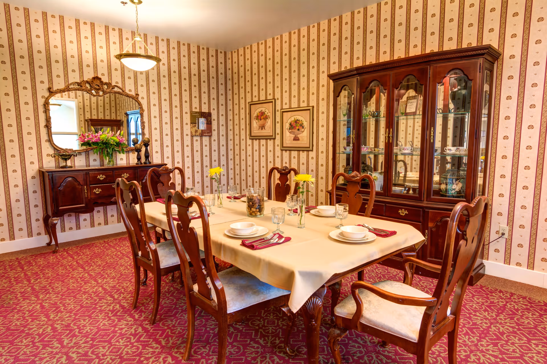 Formal dining room with a set table, wooden chairs, a china cabinet and sideboard against patterned wallpaper.