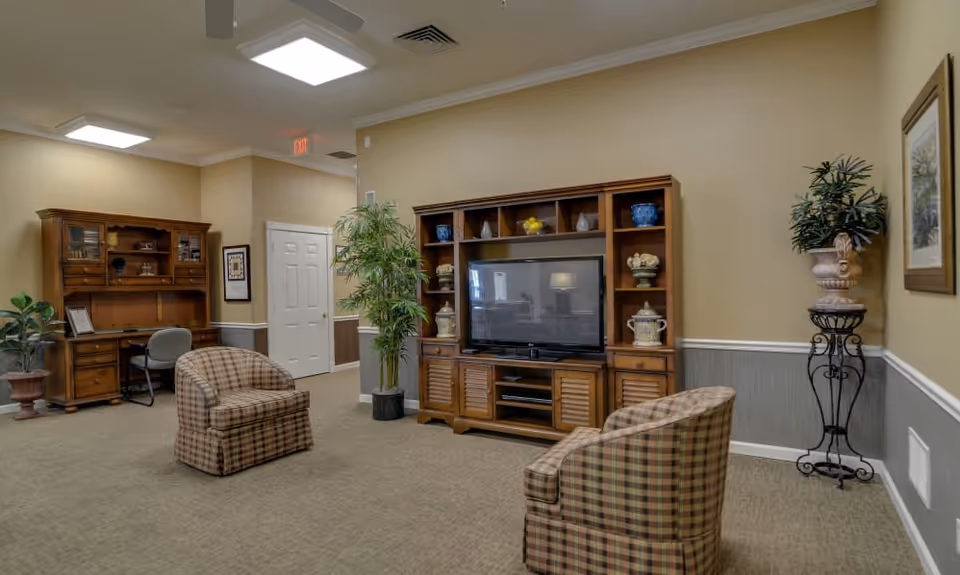 A comfortable common living room with a large TV in a wooden entertainment center, two plaid armchairs, plants, and a desk area.