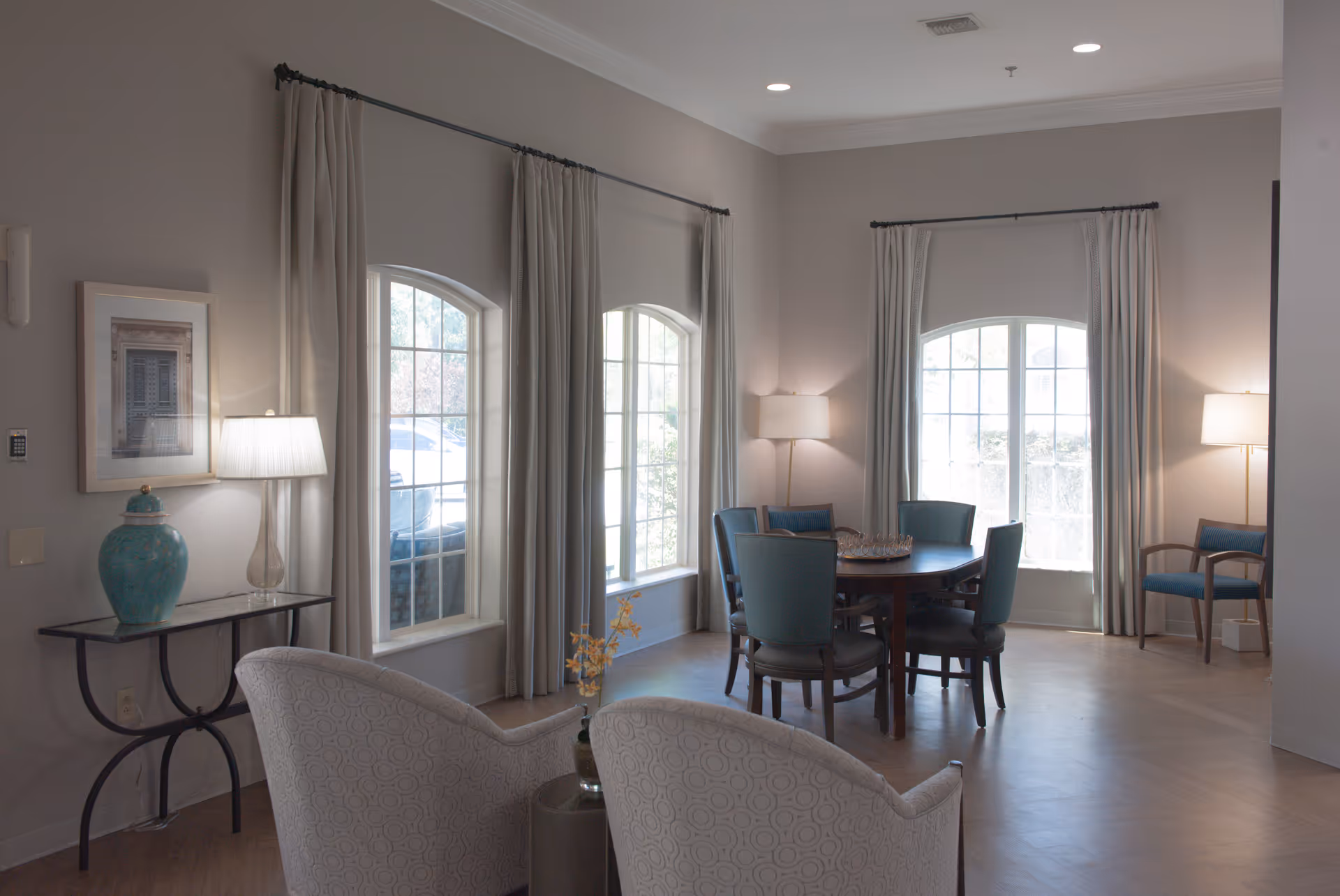 A well-lit living room area with large arched windows covered by light curtains. The room features a round wooden table surrounded by six blue upholstered chairs, two patterned armchairs in the foreground, a small side table with a blue decorative vase, and three floor lamps providing warm lighting.